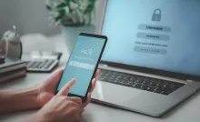 A person holds a smartphone showing a verification code in front of a laptop login screen asking for that code.