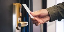 A hand holding a white hotel key card up to a gold electronic door lock.
