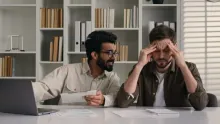 A coworker keeps talking with a clipboard in hand while the colleague beside him rubs his temples at an office desk.