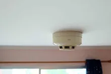 A smoke detector mounted on a white ceiling above a window in a room.
