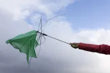 A hand holding a green umbrella turned completely inside out by the wind, its ribs exposed against a cloudy sky.