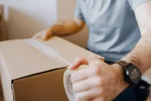A person's hand holding a roll of clear packing tape while sealing a cardboard box.