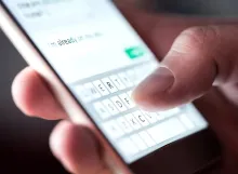 A close-up of a person's thumb typing a text message on a smartphone on-screen keyboard.