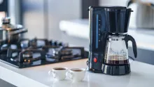 A black drip coffee maker with a nearly empty glass pot on a white kitchen counter beside a gas stove and two small cups of coffee.