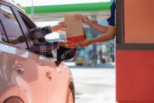A driver in a car receiving a paper takeout bag from a worker at a drive-thru window.