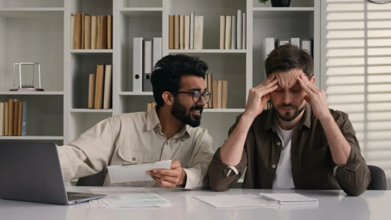 A coworker keeps talking with a clipboard in hand while the colleague beside him rubs his temples at an office desk.