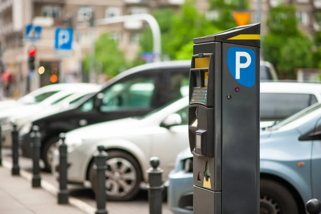 A modern parking pay station with a blue P sign in the foreground and a row of parked cars behind it on a city street.
