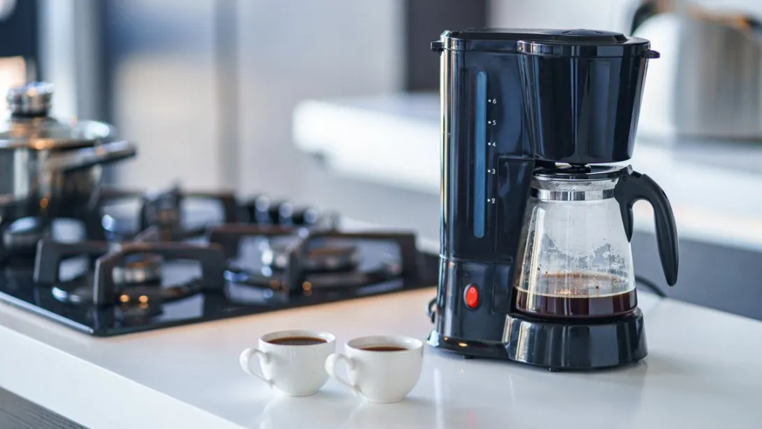 A black drip coffee maker with a nearly empty glass pot on a white kitchen counter beside a gas stove and two small cups of coffee.