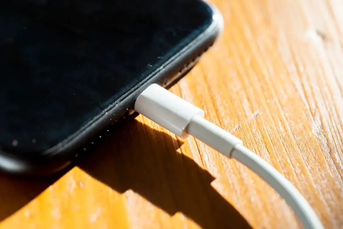 A white charging cable plugged into the bottom of a smartphone resting on a wooden table.