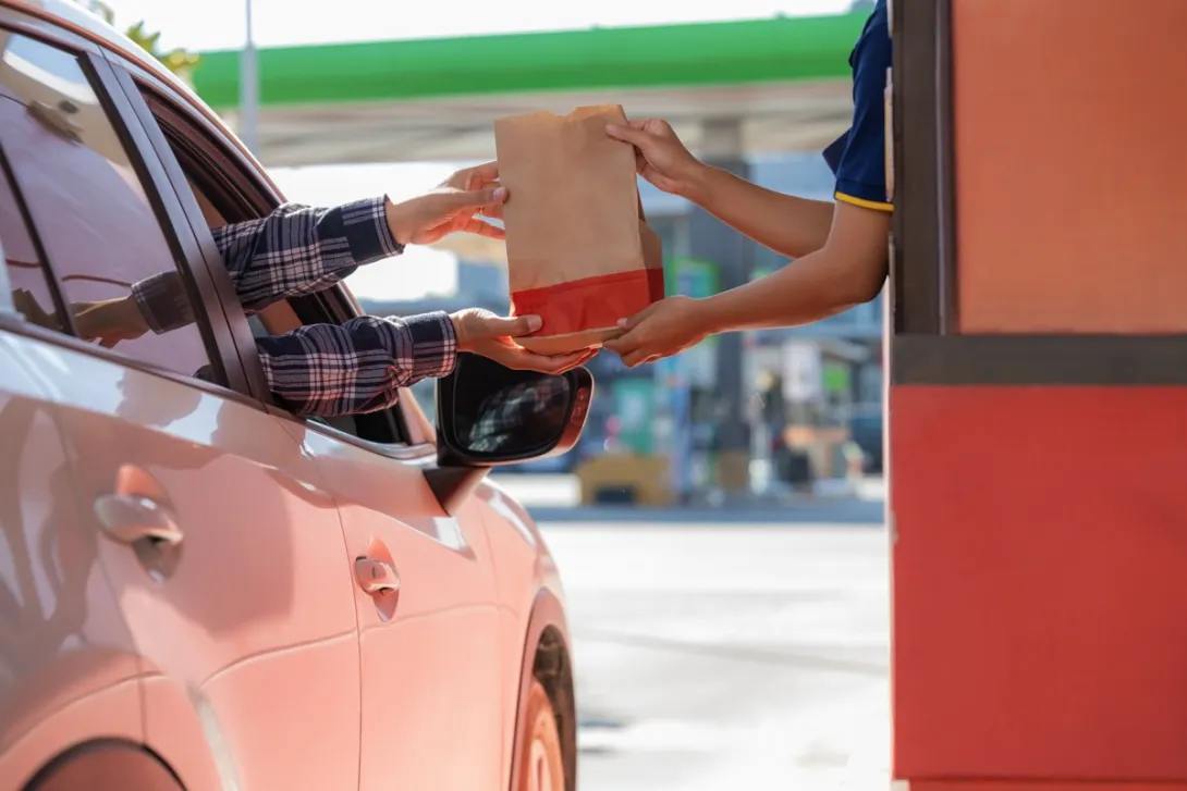 A driver in a car receiving a paper takeout bag from a worker at a drive-thru window.