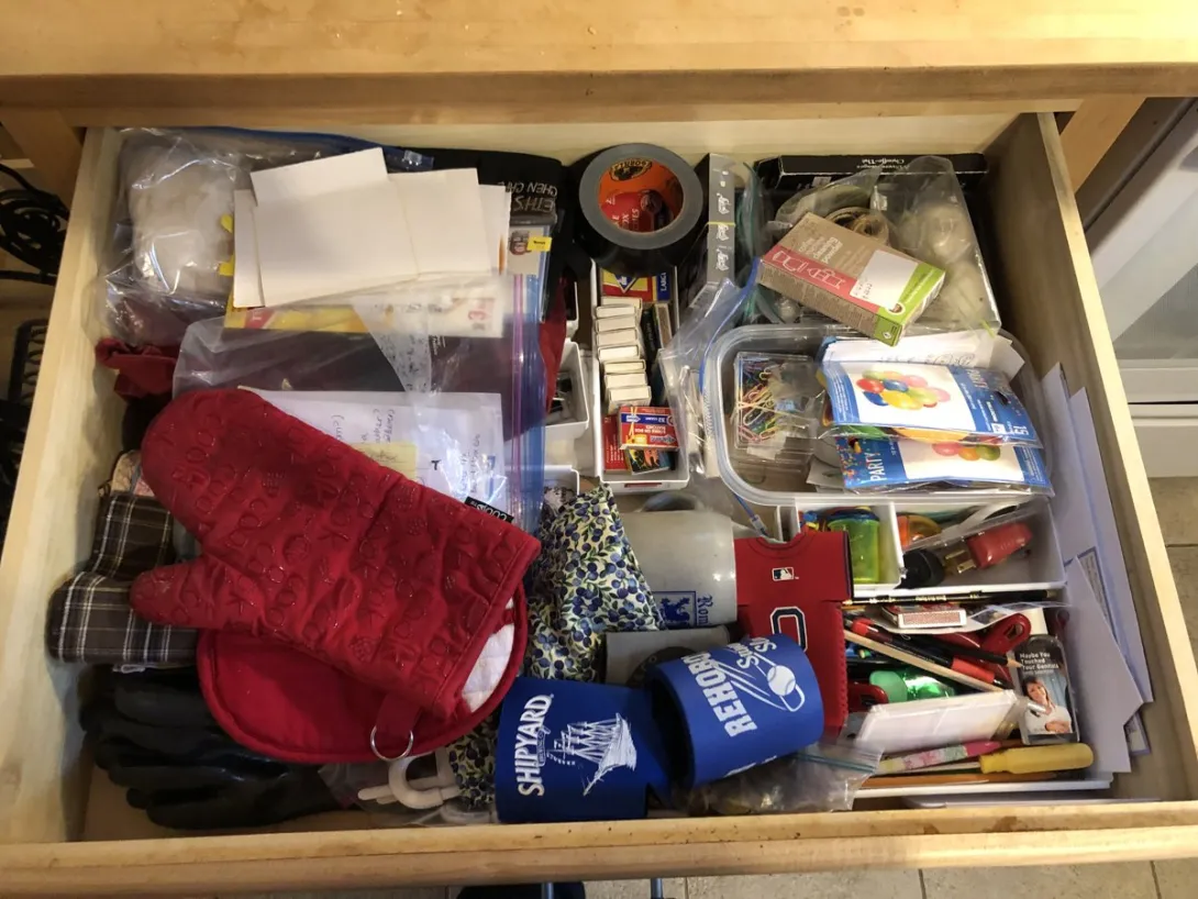An open kitchen junk drawer packed with tape, an oven mitt, tools, and assorted household clutter.