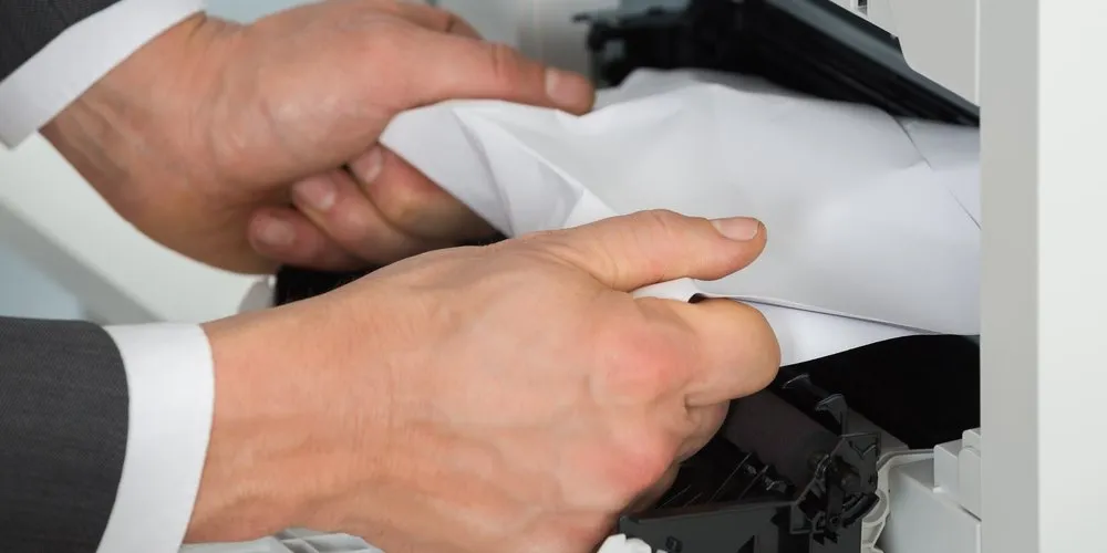 Hands pulling a crumpled sheet of jammed paper out of an office printer.